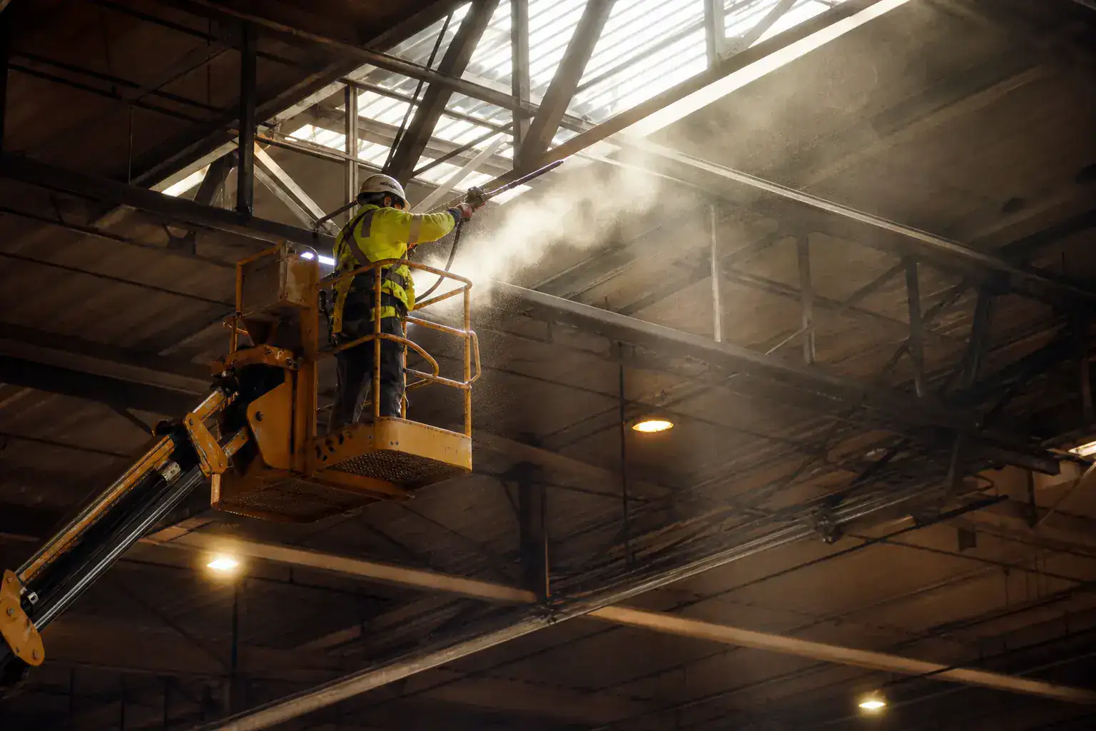 Dépoussiérage d'une charpente métallique d'atelier depuis une nacelle, faisceau de lumière dans la poussière en suspension